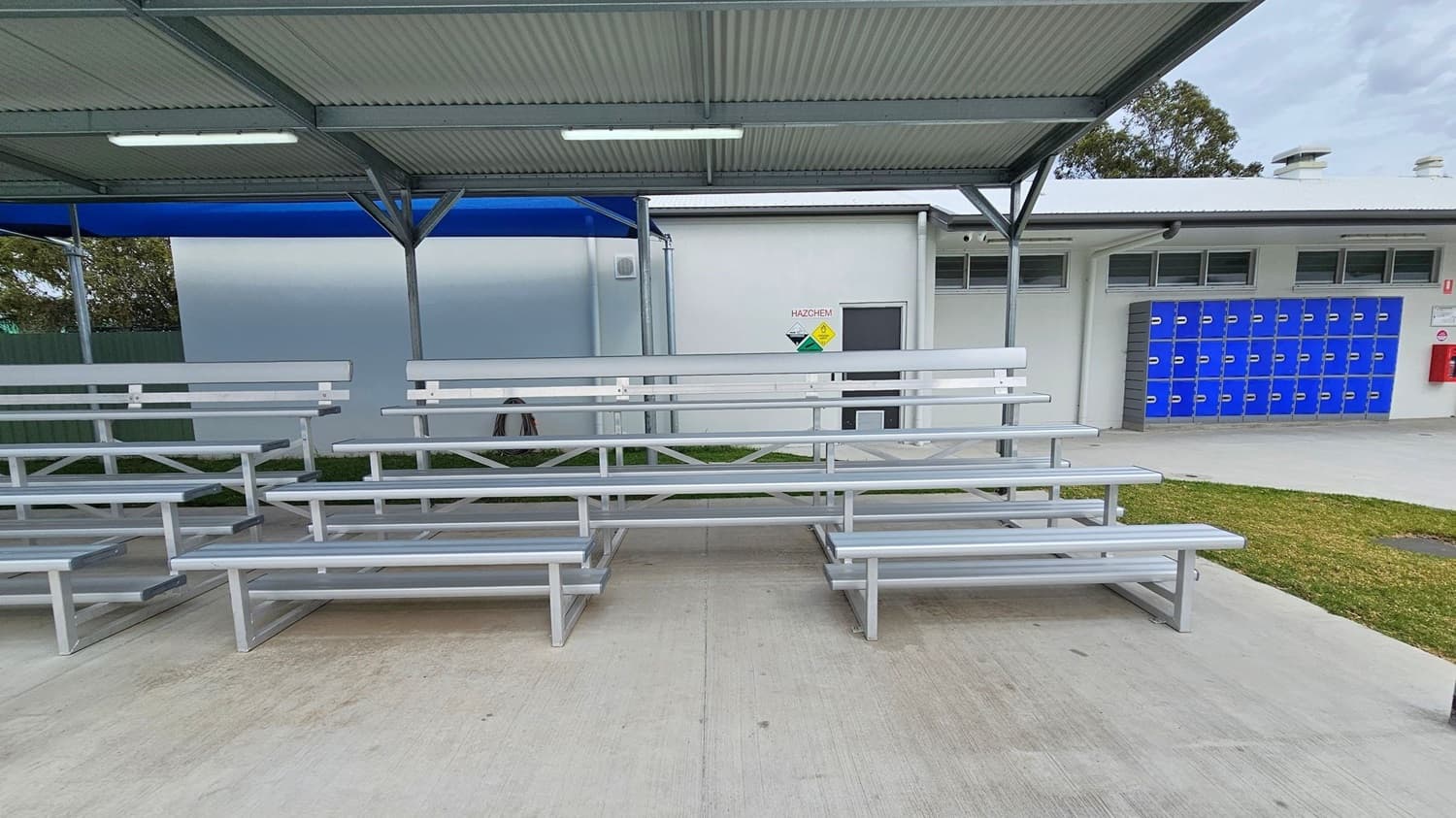 Metal bleachers are in the foreground on a concrete surface. In the background, there's a white building with a "HAZCHEM" sign and a door. To the right, a row of blue lockers is visible. The area is partially covered by a metal roof with fluorescent lights. A patch of grass is on the right side near the lockers.