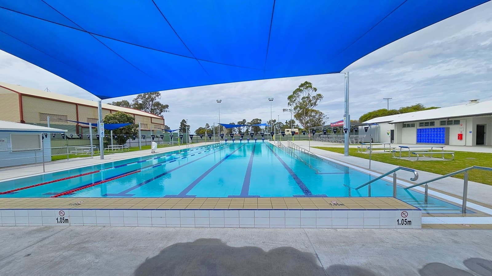 An outdoor swimming pool with clear blue water, marked with lane lines. A large blue shade cloth covers the pool area. On the left, there is a building with a green facade and a small shelter with picnic tables. On the right, a white building with blue lockers and a fire extinguisher is visible. The pool deck is concrete, and there are metal handrails leading into the water. Trees and additional structures are in the background.