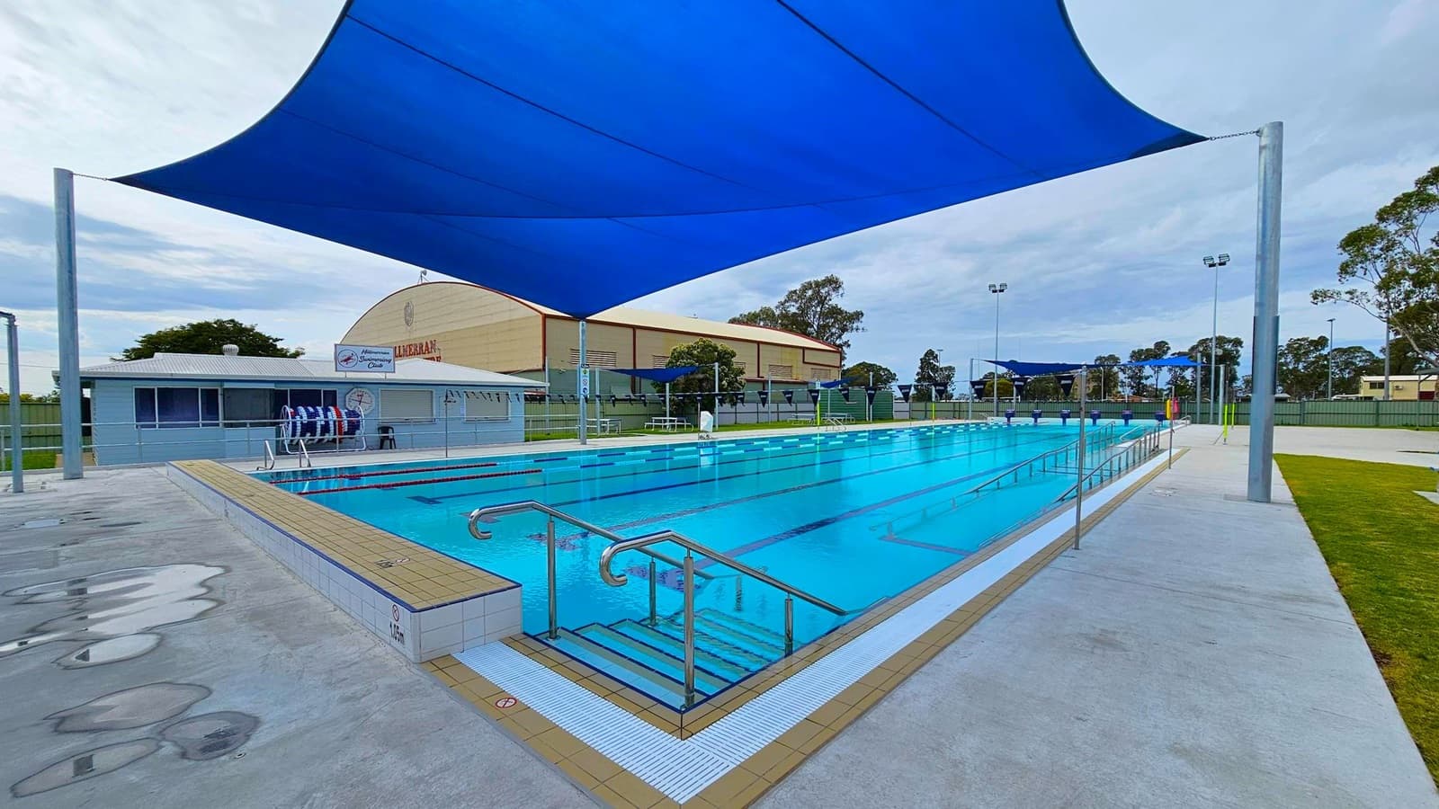 An outdoor swimming pool with clear blue water is in the centre, surrounded by a tiled deck. Metal handrails lead into the pool. A large blue shade sail is suspended above. On the left, a building with signs and pool equipment is visible. In the background, a larger building and trees can be seen. The ground is a mix of concrete and grass, and there are tall lights around the area.