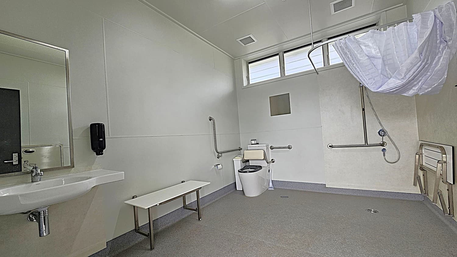A spacious, accessible bathroom with light green walls and a grey floor. On the left, there's a large mirror above a white sink with a soap dispenser mounted on the wall. In the centre, a toilet with support rails is visible. On the right, a shower area with a handheld showerhead and a curtain rail is present. A foldable bench is attached to the wall. The ceiling has ventilation panels, and there are high windows allowing natural light.