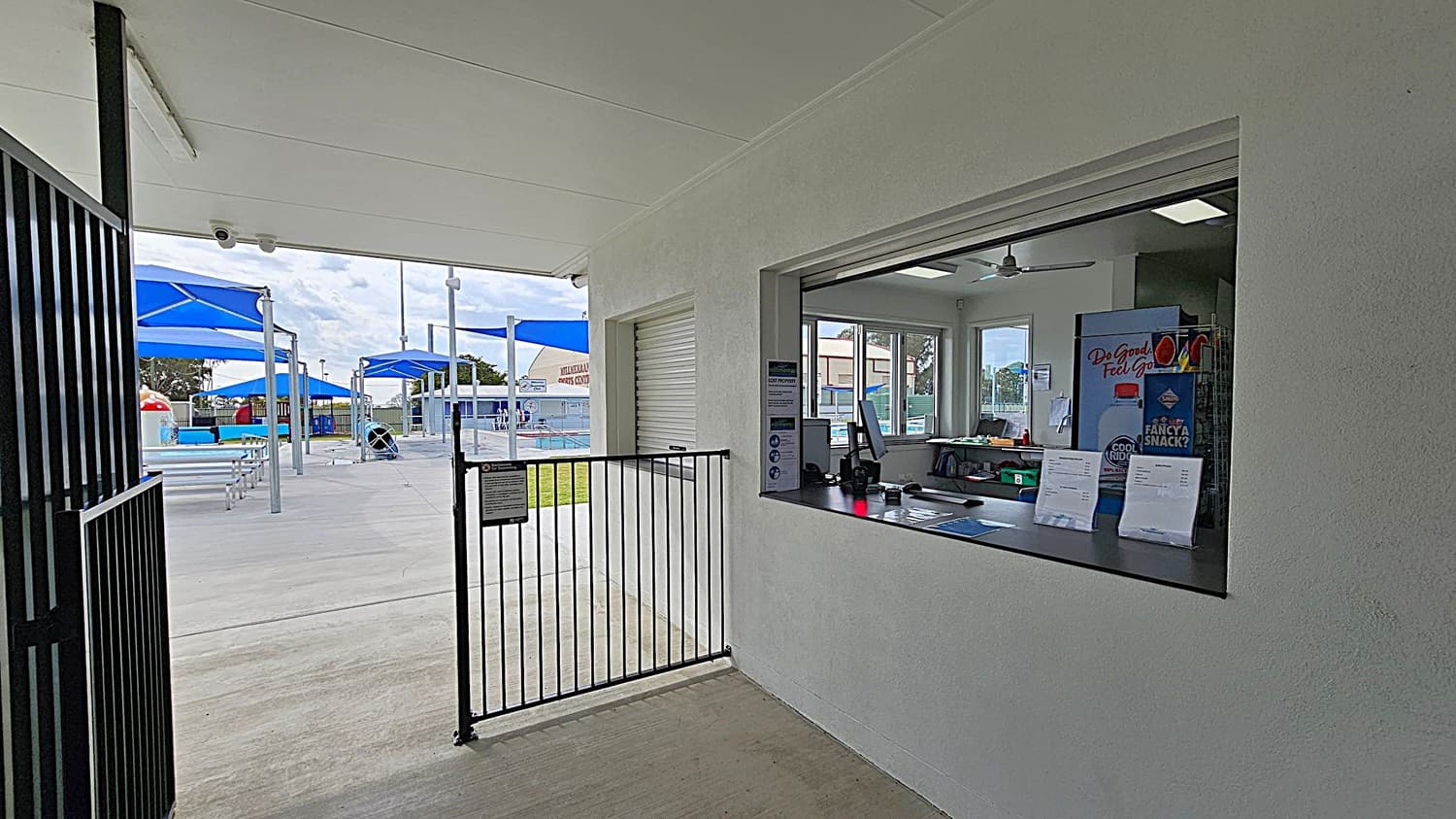 A gated entrance to an aquatic centre with black metal bars. On the right, a sign displays the facility's terms and conditions. A plaque is mounted on the wall beside it. Through the gate, blue shade structures and poolside areas are visible. The floor is concrete, and the ceiling is white with fluorescent lights. On the left, a grassy area is seen through glass panels.