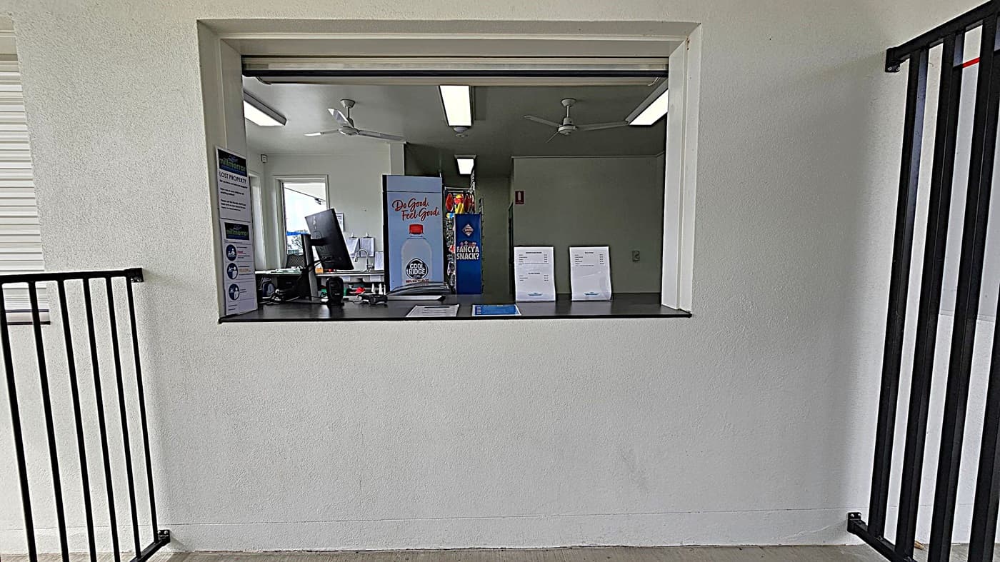 A service window set in a white wall, with a black countertop. Inside, there's a computer monitor, a "Cool Ridge" water advertisement, and a "Fancy a Snack?" sign. Two sheets of paper are displayed on the counter. The room has fluorescent lights and ceiling fans. On the left, a "Lost Property" sign is visible. Black metal railings are on both sides of the window.