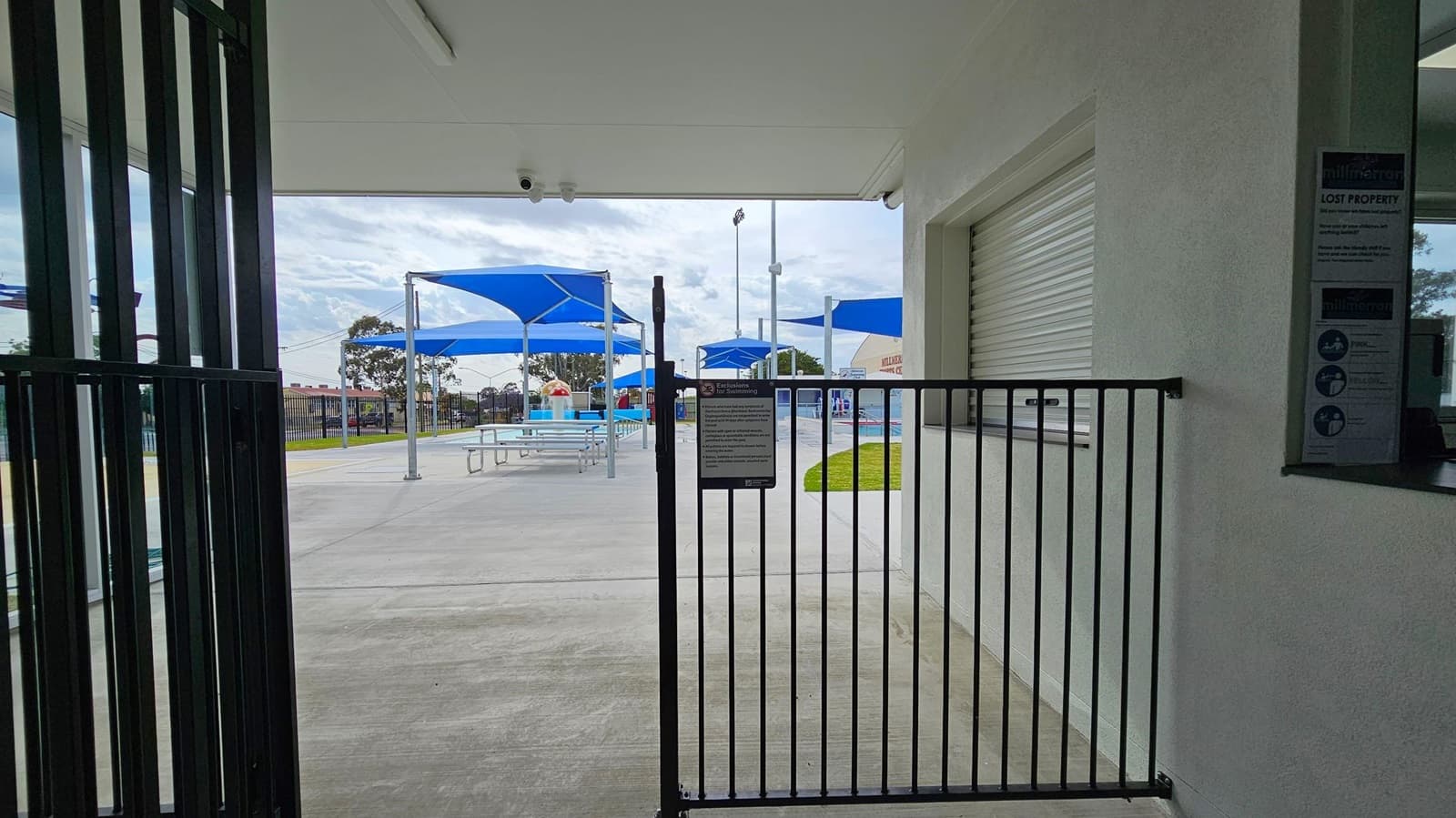 A view through a black metal gate leading to an outdoor area with blue shade structures. Under the shades are picnic tables on a concrete surface. To the right, a white wall with a closed roller shutter window and a sign. The ceiling is white with security cameras. In the background, there are trees and a fence enclosing the area.
