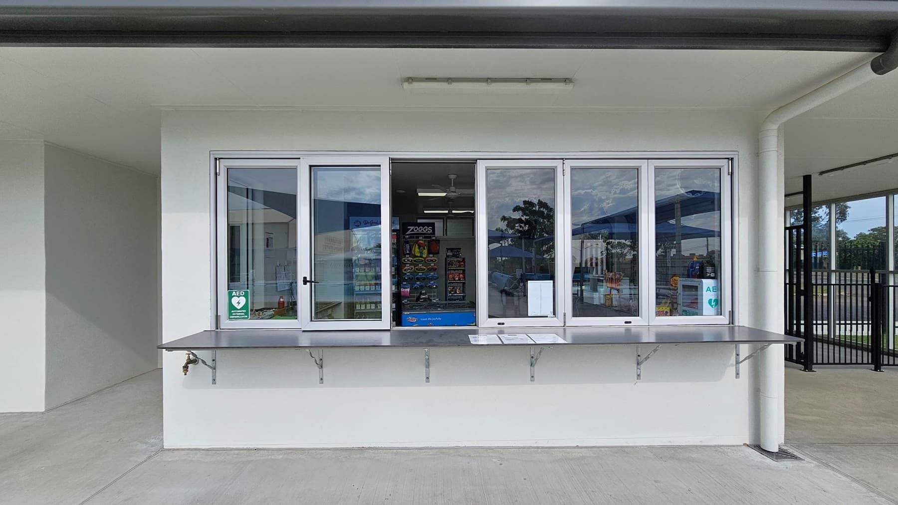 A service window with a white frame is set in a white wall. Below the window is a metal counter with brackets. Inside, there are shelves with various items, including a display for Zoggs products. A sign indicating an AED (Automated External Defibrillator) is visible on the window. The floor is concrete, and a ceiling light fixture is above the window. To the right, there is a black metal fence and a green floor area.