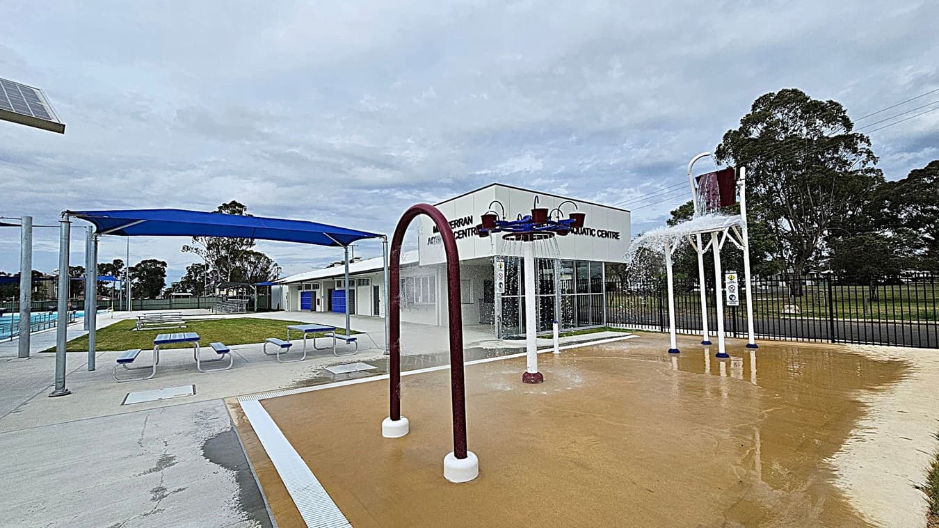 A splash pad area with water features, including a large bucket pouring water and several spray arches. To the left, there are blue shade structures over picnic tables on a concrete surface. In the background, a building with signage for an aquatic centre is visible. Trees and a fence line the perimeter, and the ground is wet from the water play area.
