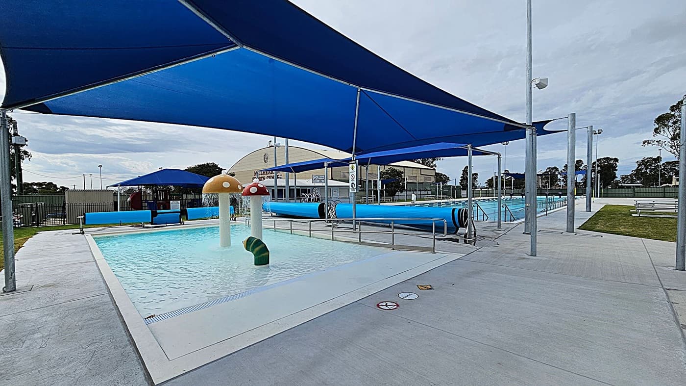 A swimming pool area with a shallow children's pool on the left, featuring mushroom-shaped water fountains. Blue shade sails cover the area, providing shelter. In the background, there is a larger swimming pool with blue pool covers rolled up on the side. Benches and a grassy area are visible on the right. A building with signage is in the background, and trees are visible beyond the fence. The ground is paved with concrete.