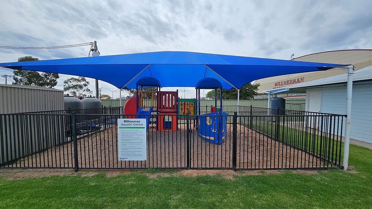 A fenced playground with a blue shade canopy overhead. Inside, there is colourful play equipment, including a slide and climbing structures. A sign in front reads "Millmerran Aquatic Centre" with information below. The ground is covered with sand, and there is grass outside the fence. In the background, a building with the word "MILLMERRAN" is visible, along with some trees and utility poles.