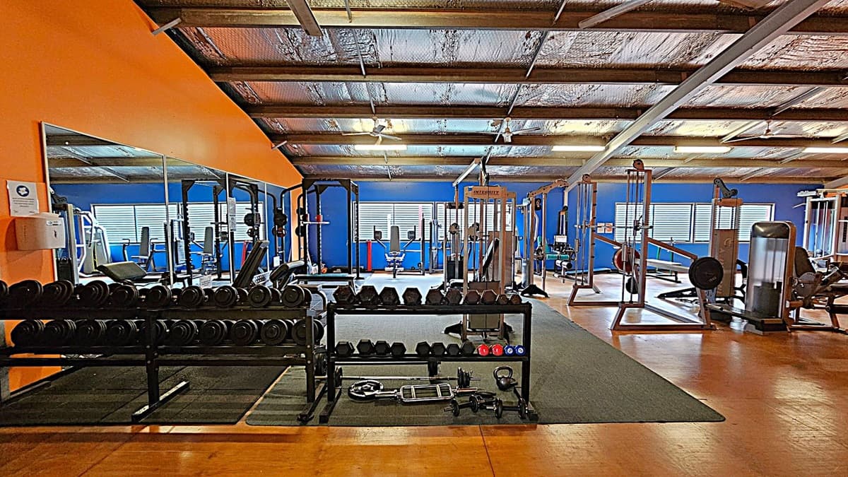 A gym entrance with a yellow door labelled "GYMNASIUM" on the left. To the right, an open doorway reveals gym equipment, including weights and machines. A whiteboard with writing and a blackboard are on the right wall. The floor is wooden, and there's a fire alarm and hand sanitiser dispenser near the door.