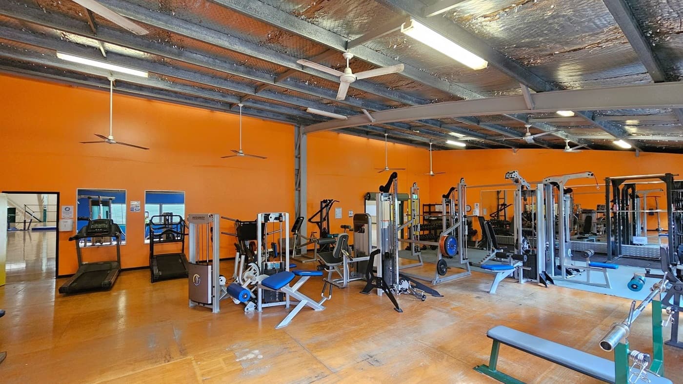 A gym with an orange wall in the background, featuring various exercise machines. On the left, there are treadmills near two windows. In the centre, there are weight machines and benches. The floor is wooden, and the ceiling is metallic with exposed beams and several ceiling fans. The lighting is provided by fluorescent lights.