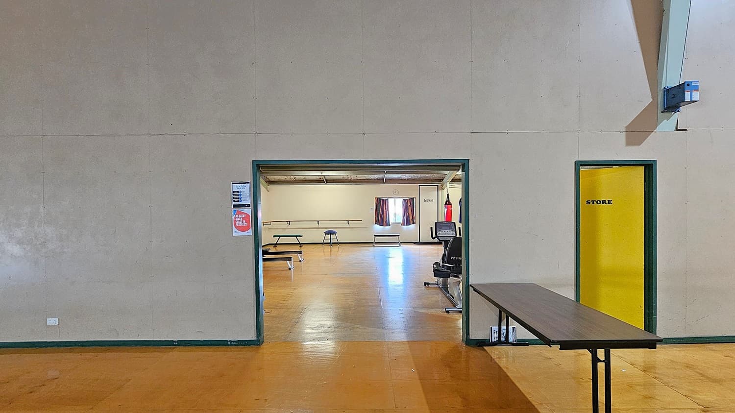 A room with a light-coloured wall and a doorway leading to another room. The floor is wooden. To the right of the doorway is a yellow door labelled "STORE". A long table is positioned against the wall on the right. Inside the adjacent room, there are tables, a stool, a punching bag, and an exercise bike. A window with curtains is visible in the background. Posters are on the wall near the doorway.