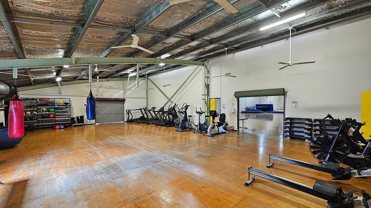 A spacious gym with a wooden floor and a high ceiling with exposed beams and insulation. On the left, there are shelves filled with various exercise balls and equipment, alongside red and blue punching bags. In the centre, several treadmills and exercise bikes are lined up against the wall. On the right, there are rowing machines and a stack of step platforms. A small open area with a table is visible through a doorway with a green roller shutter. Ceiling fans are evenly spaced throughout.