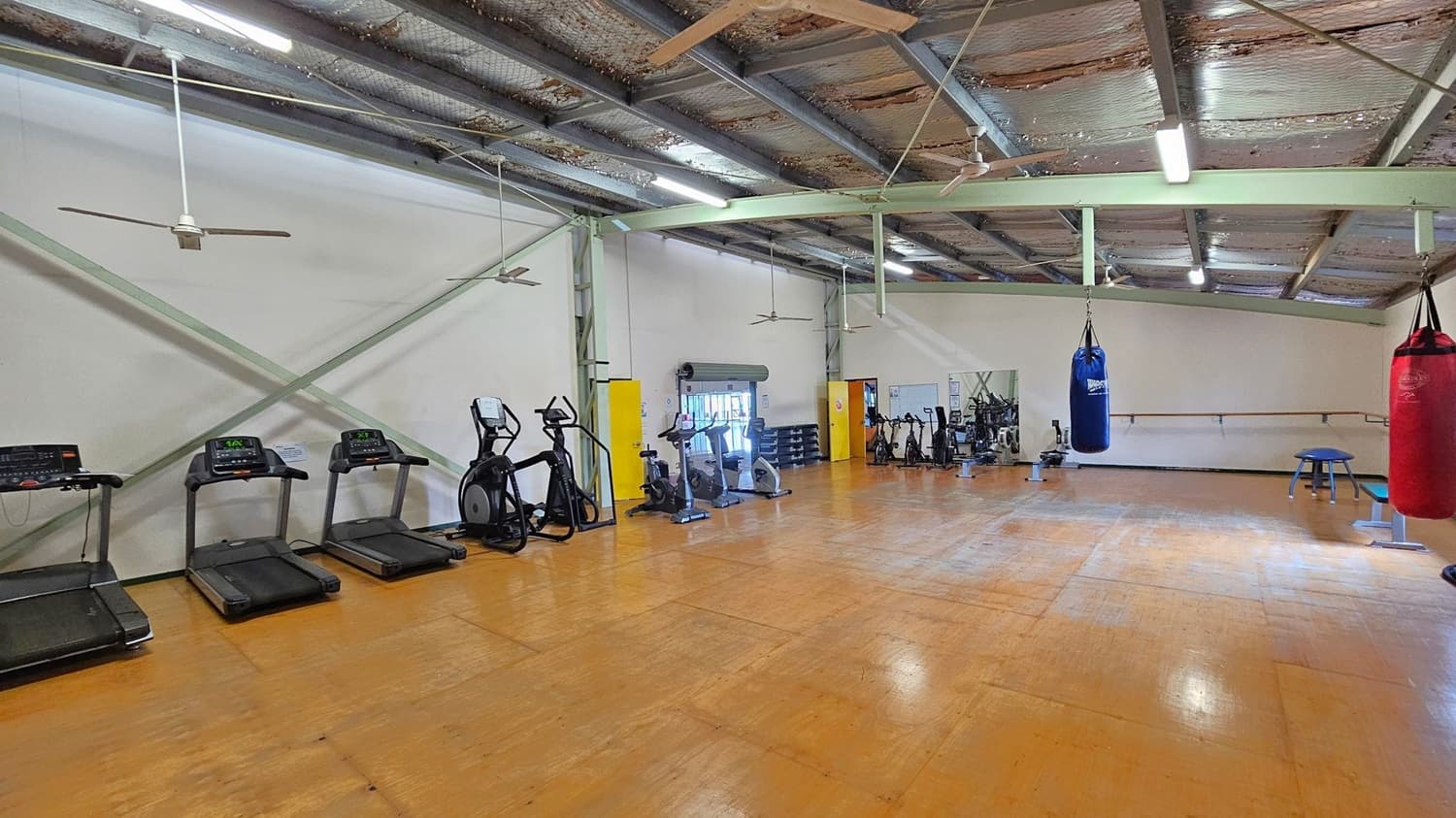 A spacious gym with a wooden floor and a high, metal ceiling. On the left, there are several treadmills and elliptical machines. In the centre, a variety of exercise bikes and a blue punching bag hang from the ceiling. On the right, a red punching bag and a small blue stool are visible. The walls are plain with a few mirrors and a ballet barre. Ceiling fans and fluorescent lights are installed above.