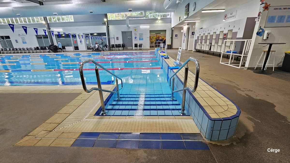 Pool stair with handrails of the 25m, 6-lane heated indoor pool. Each lane is separated by white, yellow, red, and blue floating rope lanes. At the right pool coping are white sign boards with small blue rectangular blocks to hold its position. Overhang perpendicular to the pool is a string of triangular blue flags. Against the right white wall is a white and grey cabinet locker. Above it, there is a large white sign for NO RUNNING. To the back is an open space filled with yellow life jackets.