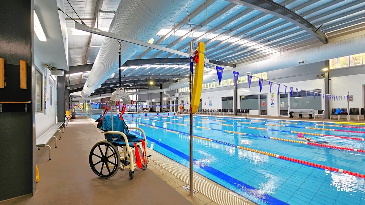 Left pool deck of 25m, 6-lane heated indoor pool with an aquatic wheelchair that has blue seating and red straps supported by a grey hoist with black rope. Found against the left white brick wall is a long, grey metal bench. On the pool coping, within the floor with a metal pole that has a yellow floating device, is a white 1.0m sign for water depth. On the bottom pool floor is the bold blue center T line that helps the swimmer stay at the center. On the walls are different precautionary signs.