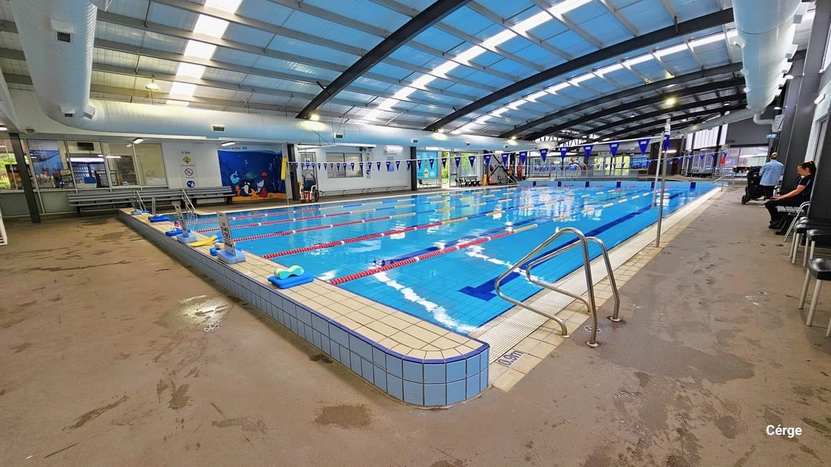 On the right pool deck of 25m, 6-lane heated indoor pool, are two stainless steel grab rails. The right pool coping has a small white sign with a blue 0.9m sign for water depth. The elevated pool coping at the start is white sign boards with small blue rectangular blocks to hold its position. Behind the long grey metal bench of the opposite pool deck is a glass window that shows a part of the café, warning signs for pool rules, and a blue mural with different sea creatures.