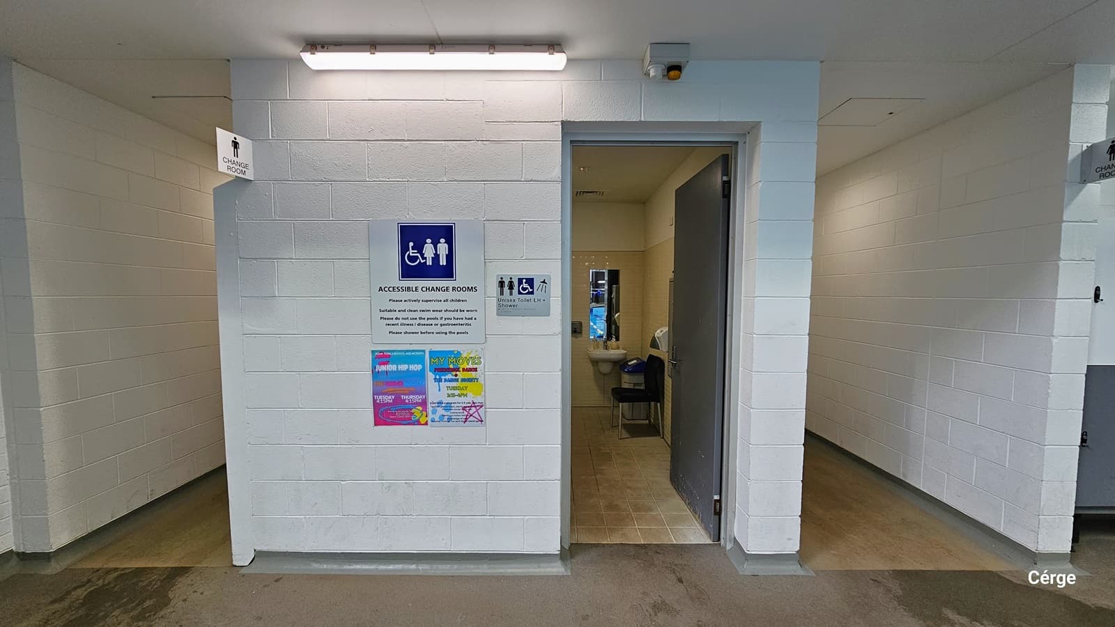 Accessible Change Room sign placed on a white brick wall at Monbulk Aquatic Center. A fluorescent ceiling illuminates the front of the change room. Underneath the Accessible Change Rooms sign are two colorful advertisements. On the right is a grey metal plate for the Unisex Toilet LH + Shower sign. Seen through the open grey metal door is the black plastic chair, wash basin with mirror, and a grey garbage bin with a blue lid. It has a beige tile wall and a brown tile floor.