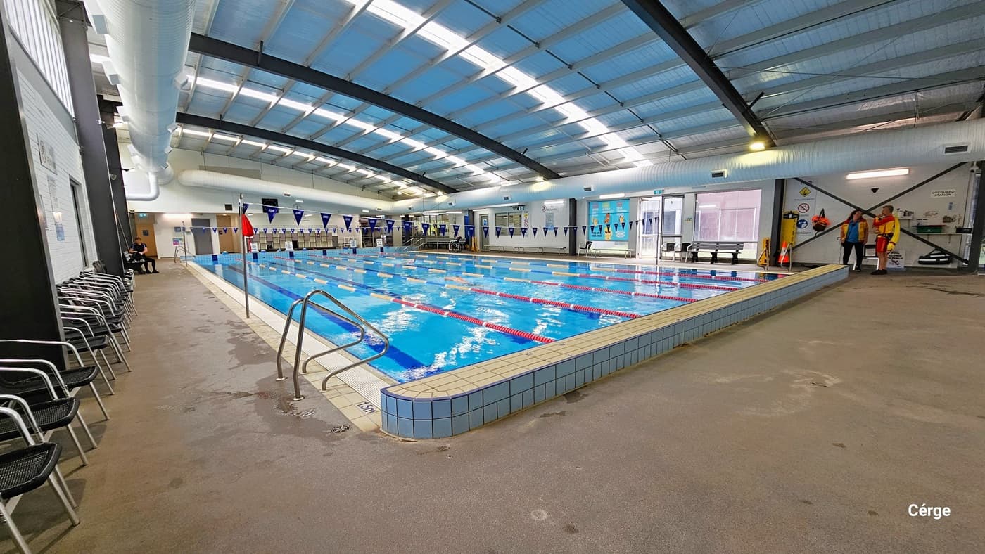 End of the 25m, 6-lane heated indoor pool are two stainless steel grab rails. On the left pool coping is a 1.5m pool depth. On the left pool deck, against the walls, are a line of black plastic chairs with stainless steel handles and legs. On the opposite pool deck are lifeguards wearing yellow and red uniforms, a yellow spine board, an orange cone, and other accessories.