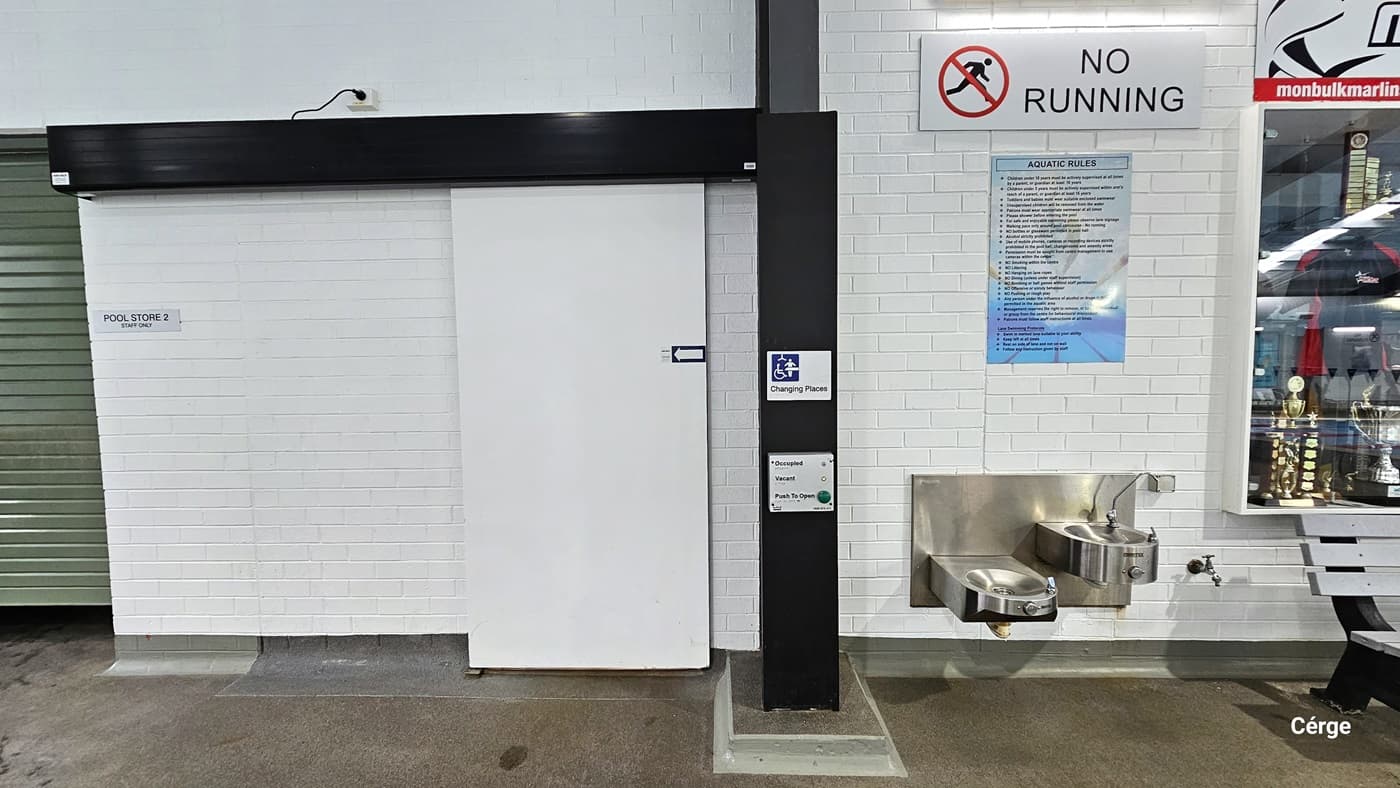 Changing Places at Monbulk Aquatic Centre has a white sliding door with an arrow pointing to the left placed within a white brick wall with black wall lining. The door can be operated through a grey metal plate that contains the green button for opening the door and two lights for occupied and vacant. On the right wall is a stainless steel, wall-mounted washbasin and drinking fountain and above it is a white NO RUNNING sign and blue aquatic rules. To the right is a glass display of trophies.
