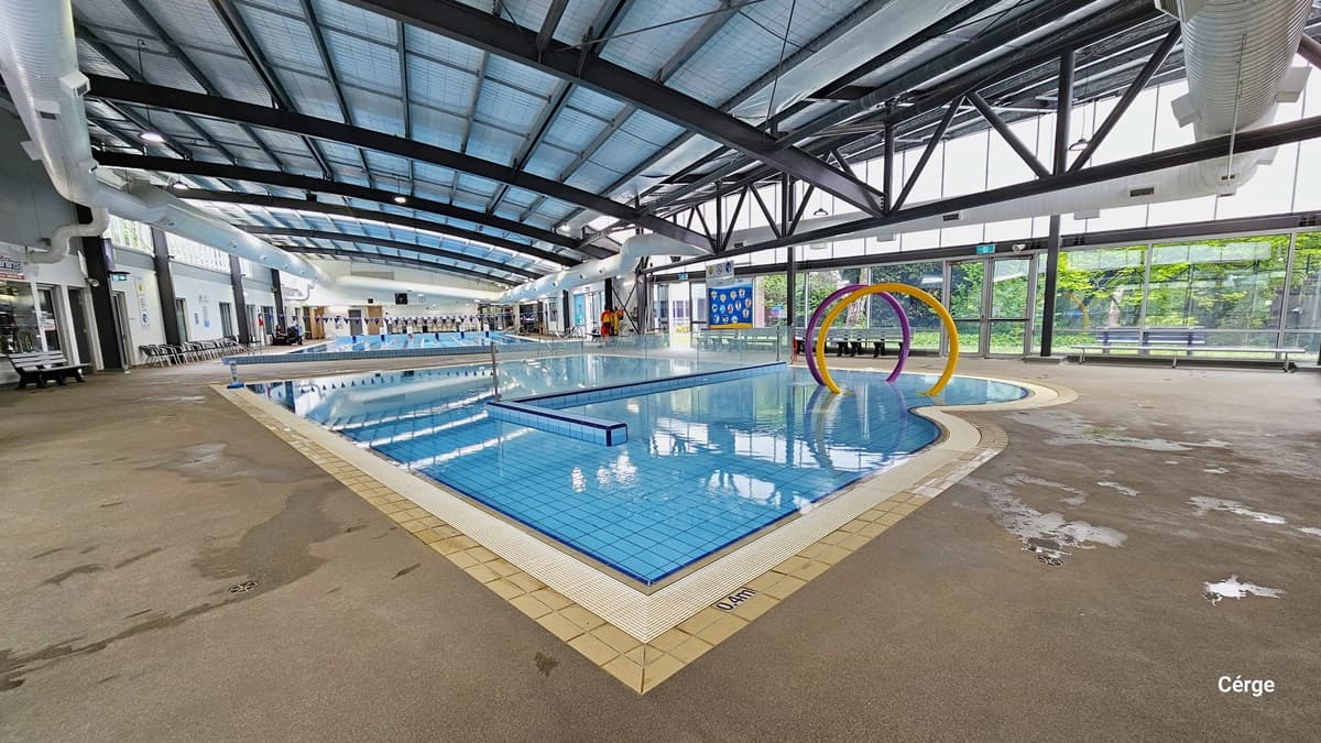 Splash water park at Monbulk Aquatic Centre has a concrete floor with rough surface, floor-to-ceiling glass windows that let natural light pass through, a white wall, and a ceiling with black metal beams and large white ventilation ducts. The splash park has a 0.4m depth detailed on the pool coping with white water grating in the middle. An L-shaped tile wall separates the pool, and it encloses the area with large yellow and purple rings.