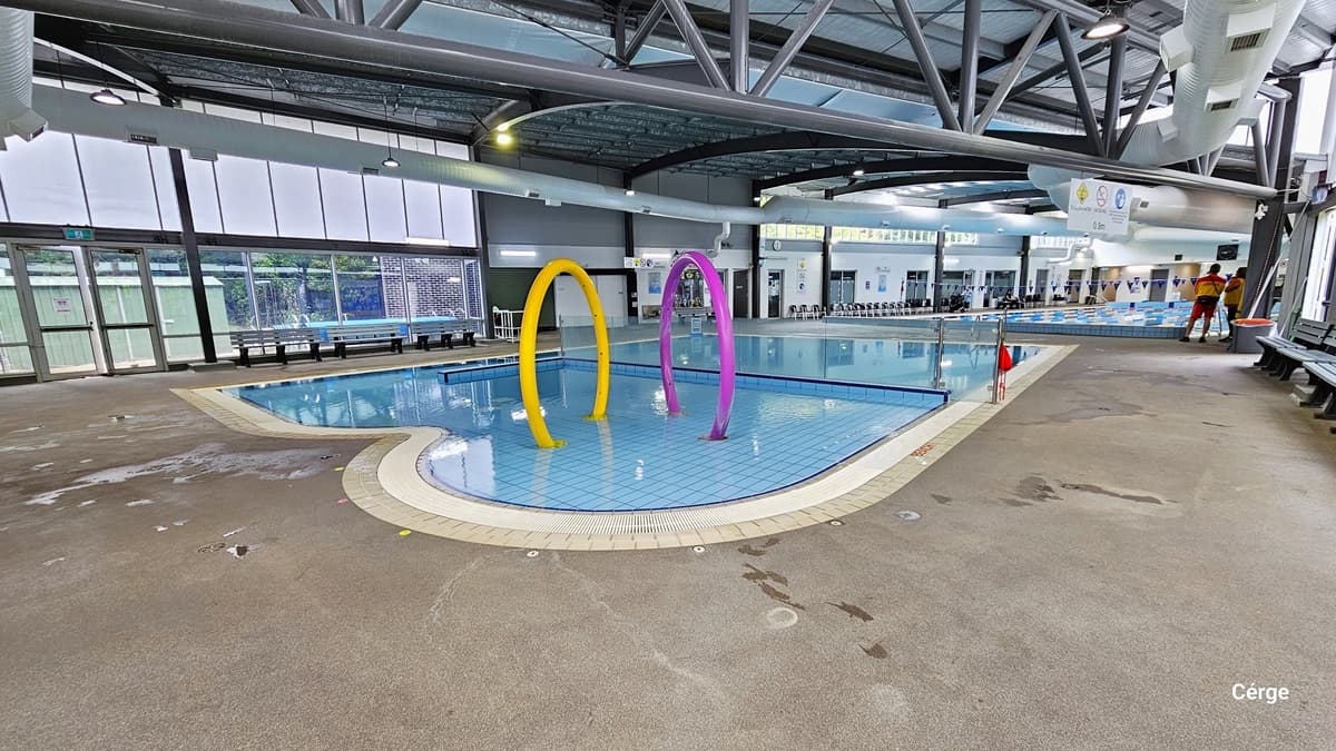 A closer look at the large yellow and purple rings within the Splash water park with 0.4m water depth. Further to the left is another floor-to-ceiling glass window with an exit glass door, which can be identified by the green exit sign. The upper glass wall is made of frosted glass while the lower is transparent. Benches and chairs can be seen all throughout the area.