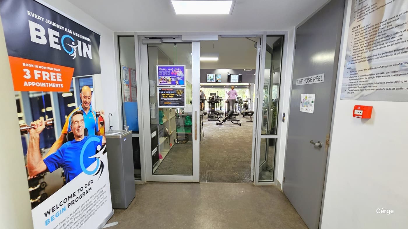 The gym entrance is through a set of double glass doors with white frames. The floor is of natural stone grey, while the walls and ceiling are white. On the ceiling just before the entrance door is a square fluorescent light. On the left side is a large poster detailing what the Begin Program is all about and a grey water fountain. On the left side is a grey door with a grey sign for FIRE HOSE REEL and a large board for facility rules. Seen through the open doors is a woman using an air walker.
