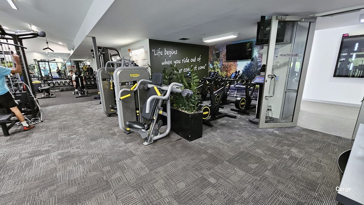 On the glass door that is open inward is a grey sign with black HEALTH CLUB letters. The door has a grey metal frame with a stainless steel U-shape handle. Behind the open glass door is a digital screen. The next section is for the black stationary bikes that have black walls with a motivational quote and a flat-screen TV mounted on the wall painted with a road and trees on the side. The other section has bench presses, dumbbells, and a lat pulldown.