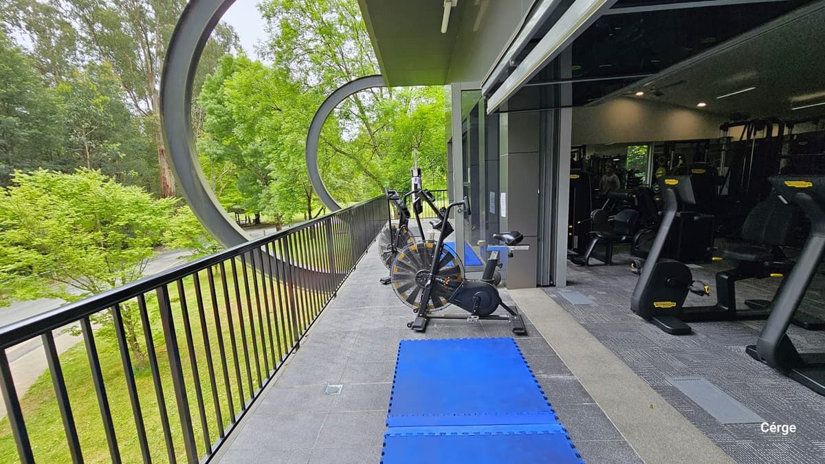 Facing the right side of the Veranda in the Gymnasium at Monbulk Aquatic Centre. The veranda has black railings and a grey tiled floor with two blue rubber mats near a stationary bike. The outside wall is grey with circular metal wall extensions. Seen through the veranda are the trees and the beautiful landscape surrounding the facility.