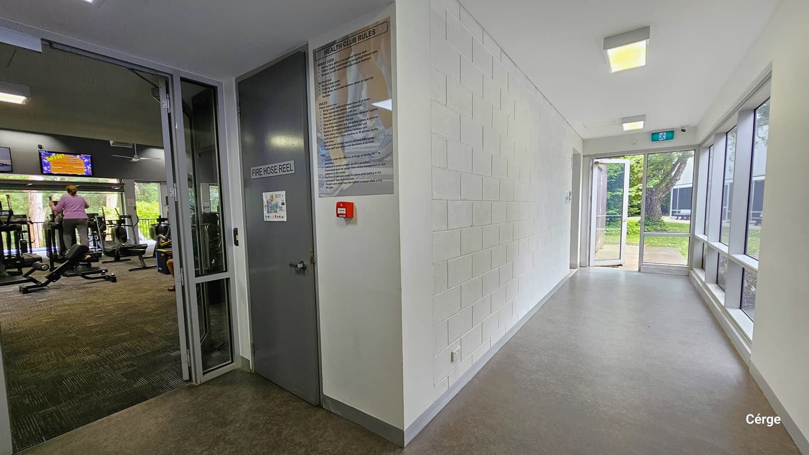 Corridor from the gymnasium leading to the exit glass doors. On the right wall from the gym entrance glass doors is a grey door with a FIRE HOSE REEL sign and a sign detailing the Health Club Rules. The floor is natural stone grey. The left wall is brick white, while the right is a combination of glass windows and white concrete wall. On the white ceiling are square fluorescent lights with warm light.