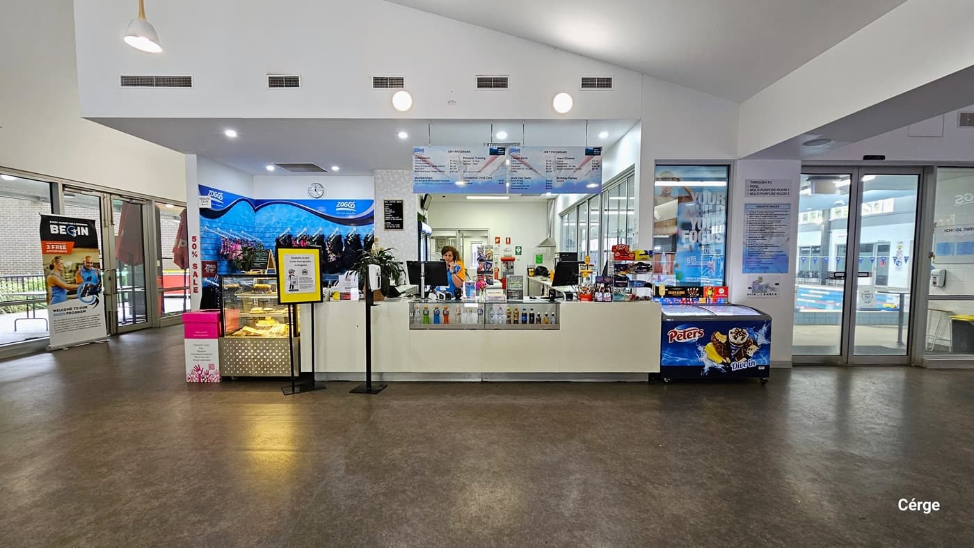 Reception desk and café inside Monbulk Aquatic Centre. The reception desk is white with a middle glass display containing varied types, sizes, and colors of drinks. In front of the desk is a white automated sanitizer dispenser placed on a short, black metal post. In front of the glass display for pastries is a small yellow banner for the Cergé companion app. On the blue wall section behind the glass display are different sorts of swimwear.
