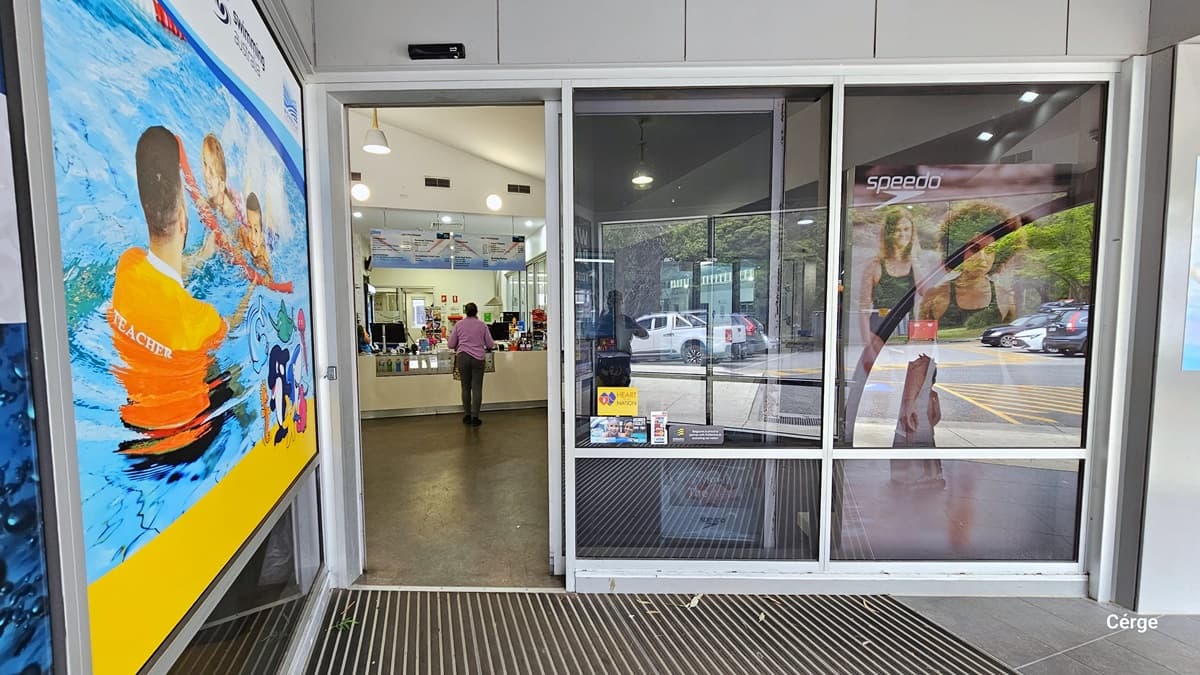 Automatic, glass sliding door entrance with white metal frames to Monbulk Aquatic Centre. On the left side of the entrance door is a large poster with an image of a male swimming coach wearing an orange shirt with the word TEACHER at the back of the shirt in white, guiding two children in the pool. Seen through the open door is a woman standing in front of a reception desk. The outside walls are gray, and the entrance floor has a stainless steel grating, while inside, is a white wall.