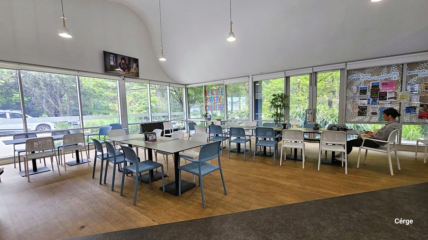 Seating area of the café. The floor space is a combination of brown and grey flooring. There are white square tables with black metal leg support and white and sky blue netted chairs. Near the right corner glass window is a red and blue ‘BE ACTIVE 6 WEEK CHALLENGE’ in capital letters. There is also a brown-and-white print bulletin board for What’s on in Our Community with photos and infographic materials. Mounted on the white wall, right above the glass window, is a flatscreen TV.