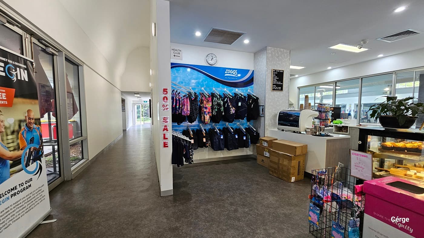A closer look at the swimwear and swimming accessories of the reception desk and café inside Monbulk Aquatic Centre. The blue wall display at the back with the white ZOGGS logo showcases upper and lower swimwear for kids. Above it is a white clock, a white Health Club sign with an arrow pointing to the left passageway that has a large white gym advertisement at its entryway. Detailed on the white wall that separates the passageway and the café is a red 50% SALE.