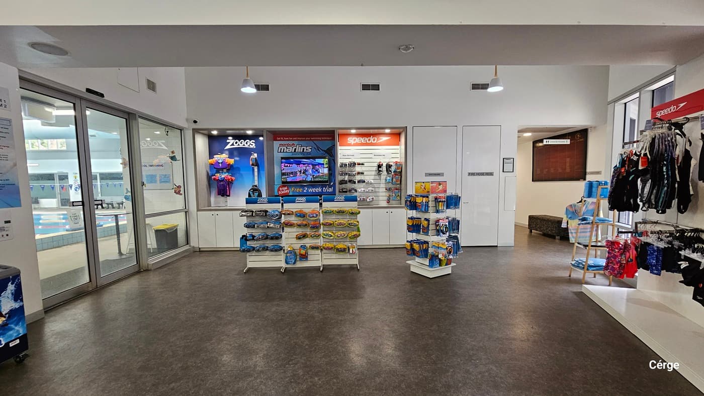 A display of swimwear and swimming accessories close to the entrance of the indoor pool accessible on the right of the reception and café. Mounted on the wall are upper and lower swimwear white on the white racks are swimming accessories such as goggles and swim caps. To the back are two white doors with a grey WHITE HOSE REEL sign next to a corner with a grey bench and a large, red glass wall display. The floor is natural stone grey, and the walls are white.