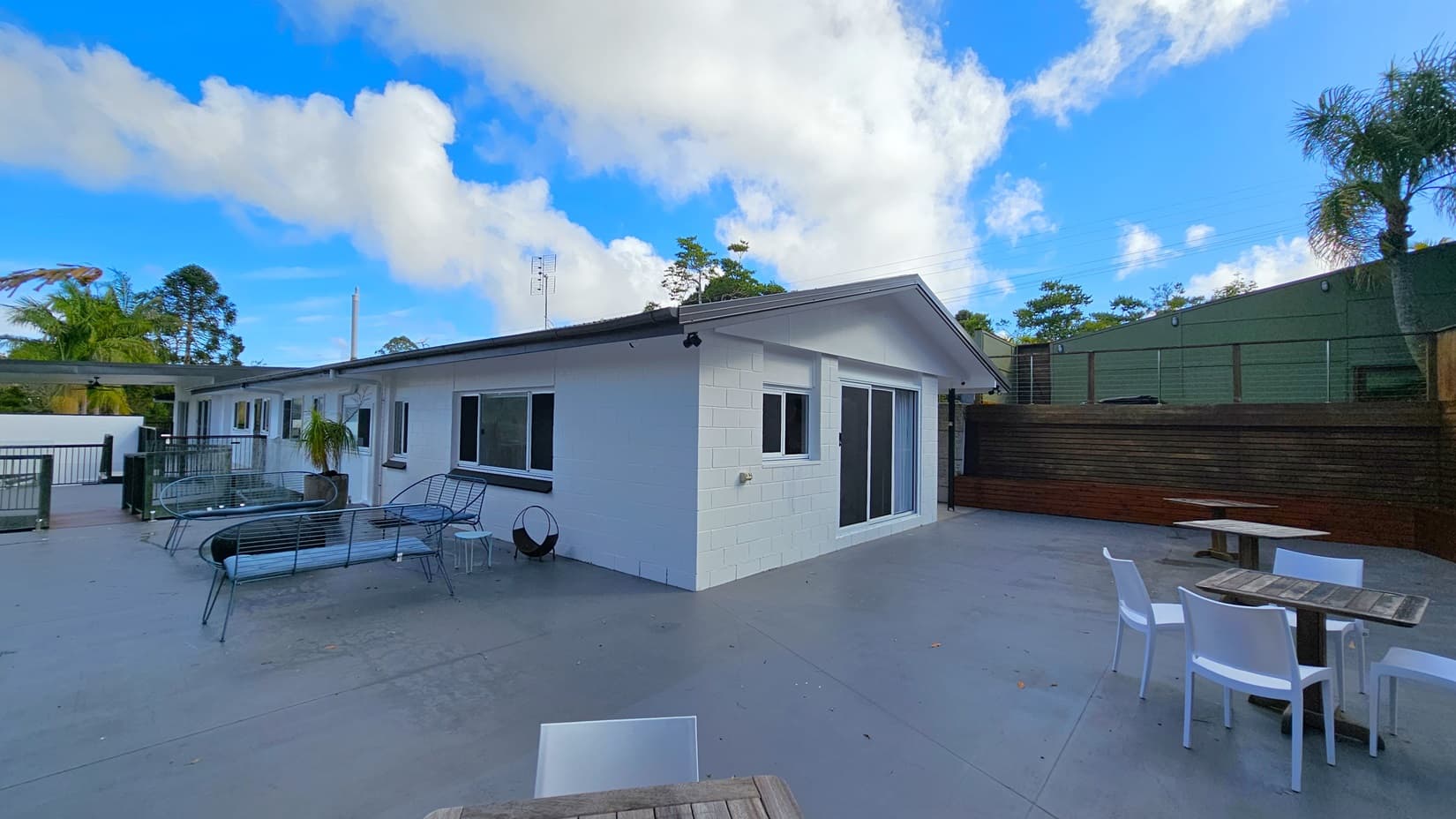 On the left, there is a white building with multiple windows and a sliding glass door, accompanied by black metal benches and a small table. In the middle, a spacious grey concrete patio extends across the area. On the right, there are wooden tables with white plastic chairs, and a tall wooden fence with greenery behind it. The background features tall trees and dense foliage.