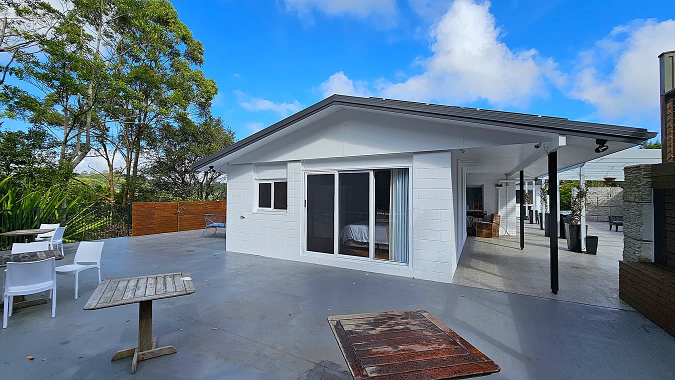 On the left, there are white plastic chairs and tables on a grey concrete patio, with tall trees and a wooden fence in the background. In the middle, a white building with a sliding glass door and a small window is visible. On the right, the patio continues with black metal columns supporting a white overhang, leading to another section of the building. The floor is grey concrete, and the ceiling of the overhang is white with recessed lighting.