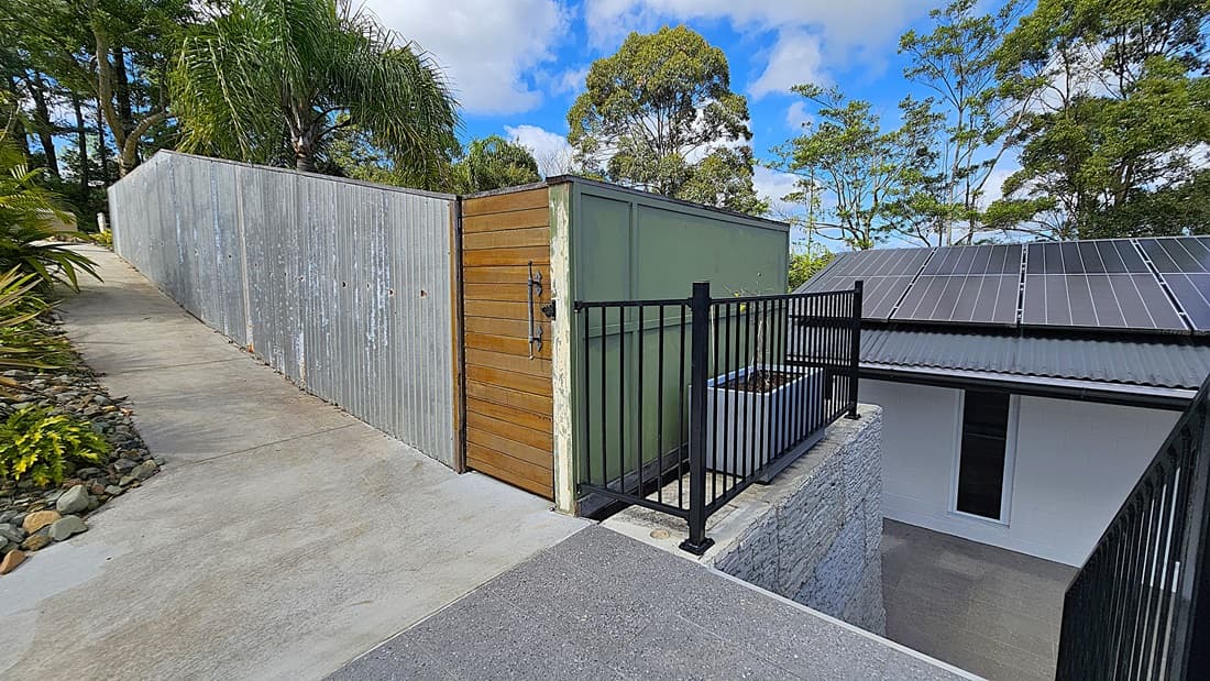 On the left, a concrete pathway bordered by rocks and plants leads uphill alongside a corrugated metal fence. In the middle, a wooden gate with a metal handle is attached to a green structure. On the right, a black metal railing surrounds a planter box and overlooks a white building with a grey roof fitted with solar panels. The background features tall trees and dense foliage.