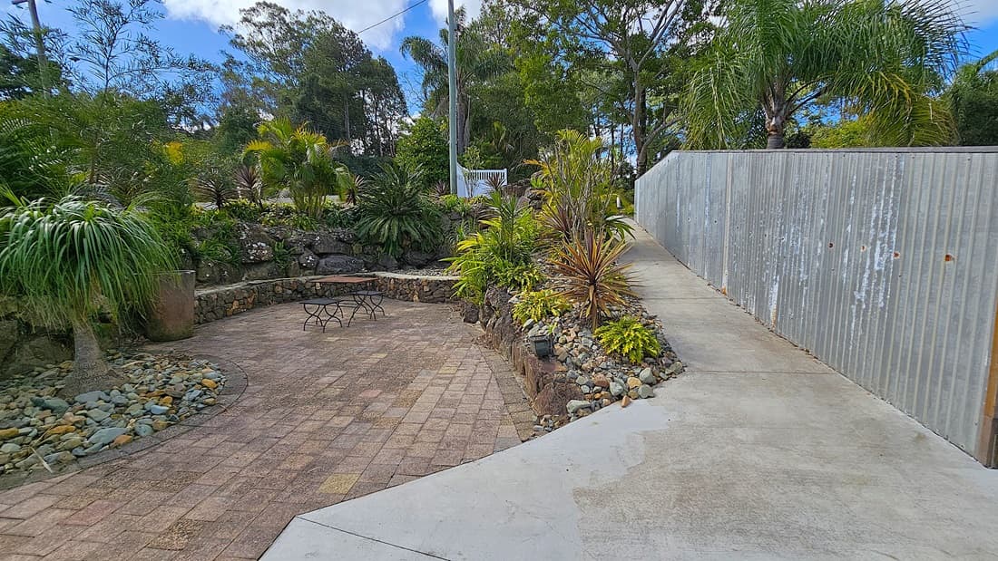 On the left, there is a garden area with various plants and a large potted plant, bordered by a stone wall. In the middle, a circular paved area with a small metal table and chairs is surrounded by more plants and rocks. On the right, a concrete pathway runs alongside a corrugated metal fence, leading uphill. The background features tall trees and dense foliage. The floor is a mix of concrete and paving stones.