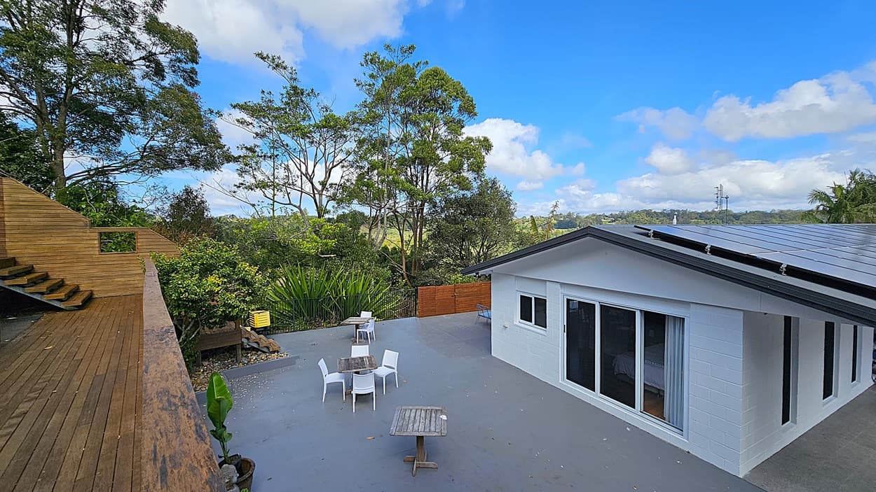 On the left, a wooden staircase ascends alongside a wooden deck bordered by lush greenery. In the middle, a grey concrete patio features several white plastic chairs and wooden tables. On the right, a white building with large sliding glass doors and a grey roof with solar panels is visible. The background consists of tall trees and dense foliage.