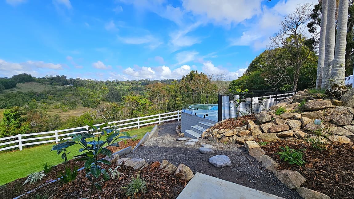 On the left, a white wooden fence runs along a green lawn with various plants and shrubs. In the middle, a gravel pathway with stepping stones leads to a raised deck with a round above-ground pool. On the right, a stone retaining wall supports a garden area with large rocks and tall palm trees. The background features rolling hills and dense forest. The floor is a mix of gravel, stepping stones, and grass.