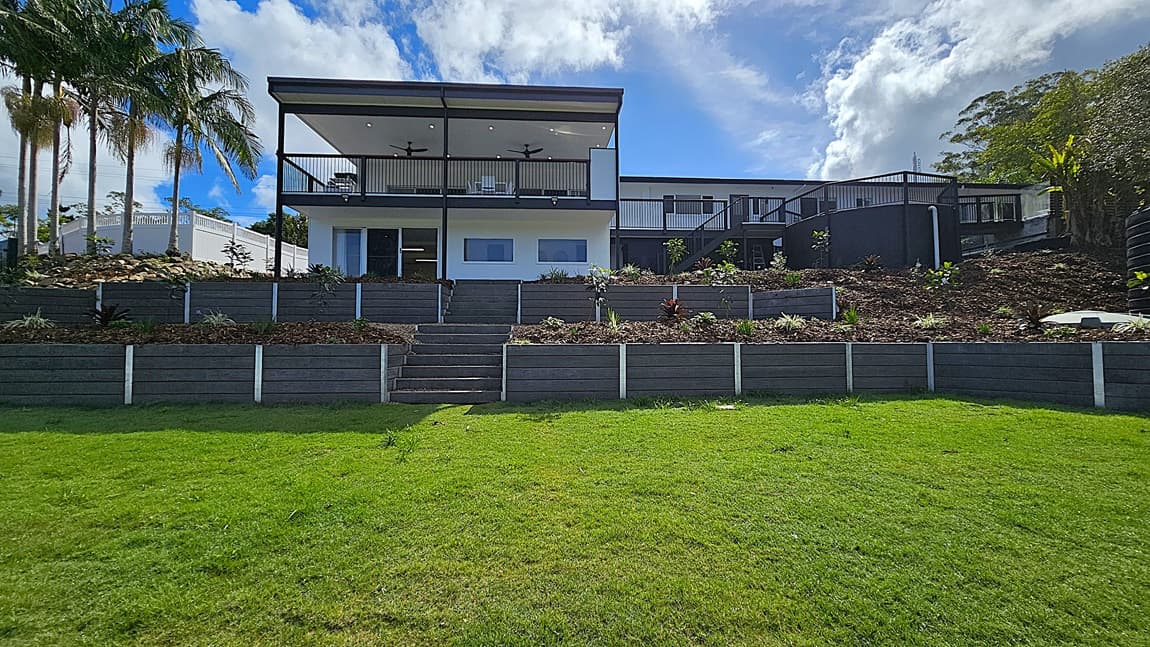 On the left, tall palm trees stand next to a white building with a sloped roof. In the middle, a modern two-storey house with a large balcony supported by black columns overlooks a tiered garden with stone steps leading up to it. On the right, the house extends with additional rooms and a black staircase. The background features dense foliage and more trees. The floor is a mix of green grass and stone steps, and the ceiling of the balcony is white with recessed lighting and ceiling fans.