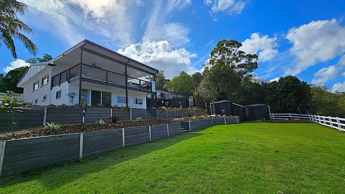 On the left, a modern two-storey white house with a large balcony supported by black columns. In the middle, tiered garden beds with various plants and a stone pathway leading up to the house. On the right, two large black water tanks are situated near a white wooden fence. The background features dense trees and foliage. The floor is a mix of green grass and stone pathways, and the ceiling of the balcony is white with recessed lighting and ceiling fans.