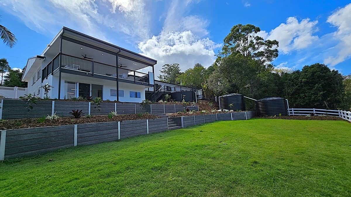 On the left, a modern two-storey white house with a large balcony supported by black columns. In the middle, tiered garden beds with various plants and a stone pathway leading up to the house. On the right, two large black water tanks are situated near a white wooden fence. The background features dense trees and foliage. The floor is a mix of green grass and stone pathways, and the ceiling of the balcony is white with recessed lighting and ceiling fans.