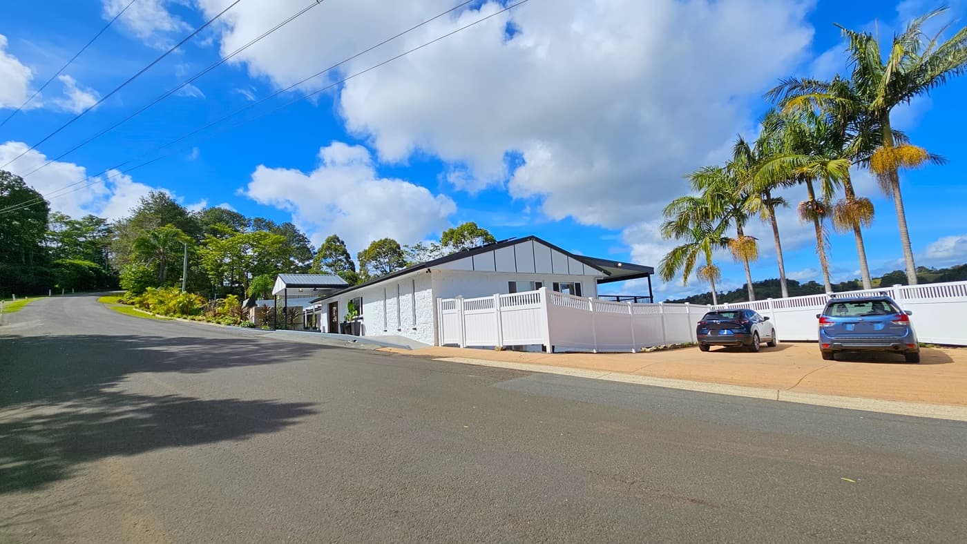 On the left, there is a sloped asphalt road bordered by a grassy verge with various plants and trees. In the middle, a white building with a black roof and white fence runs parallel to the road. On the right, two parked cars are situated on a paved area next to the building, with tall palm trees behind them. The floor is a mix of asphalt and paving. The ceiling is a black gabled roof extending over the building. The background features dense greenery and additional trees.