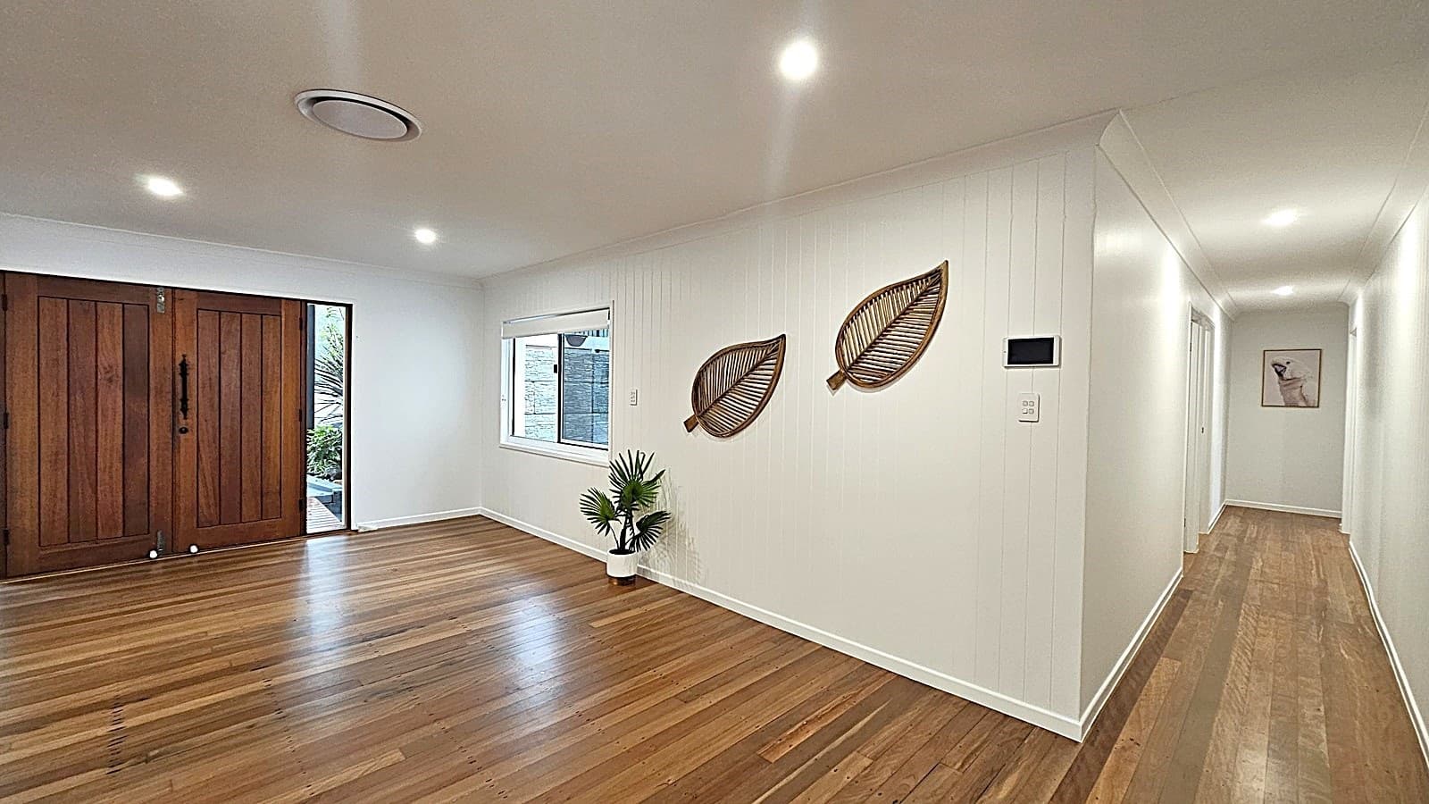 On the left, there are large wooden double doors with black handles, next to a window with a white frame. In the middle, a white wall features two decorative leaf-shaped wall hangings and a small potted plant on the floor. On the right, a long hallway extends into the distance, with a framed picture on the far wall. The floor is made of polished wooden planks, and the ceiling is white with recessed lighting and a circular air vent.