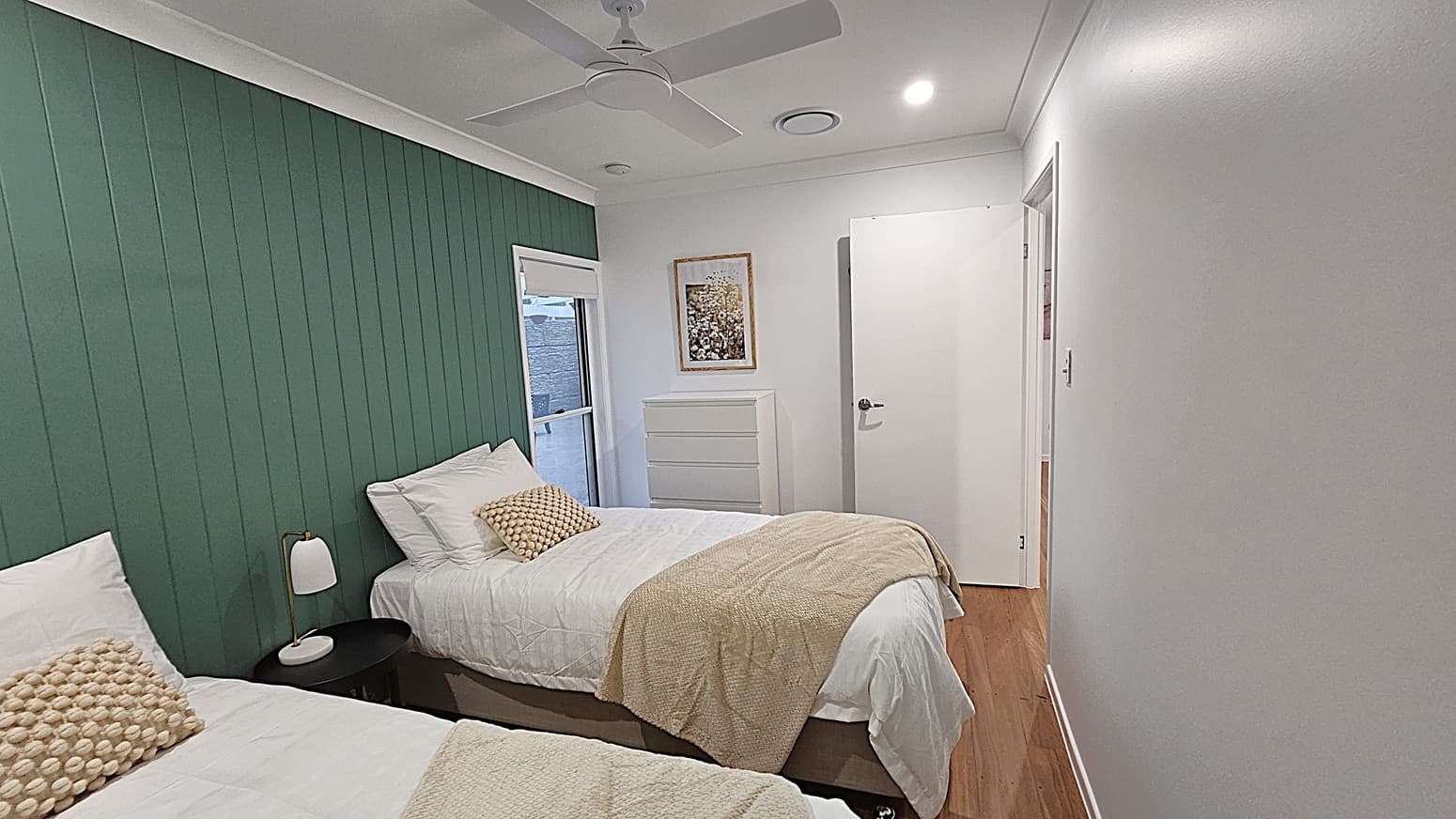 On the left, there is a wooden front door with glass panels and a window with a white blind. A potted plant sits on the floor near the corner. In the middle, two decorative wooden leaf wall hangings are mounted on a white wall. On the right, a long hallway with wooden flooring leads to a distant wall with a framed picture. The floor is polished wooden planks, and the ceiling is white with recessed lighting and a circular air vent.