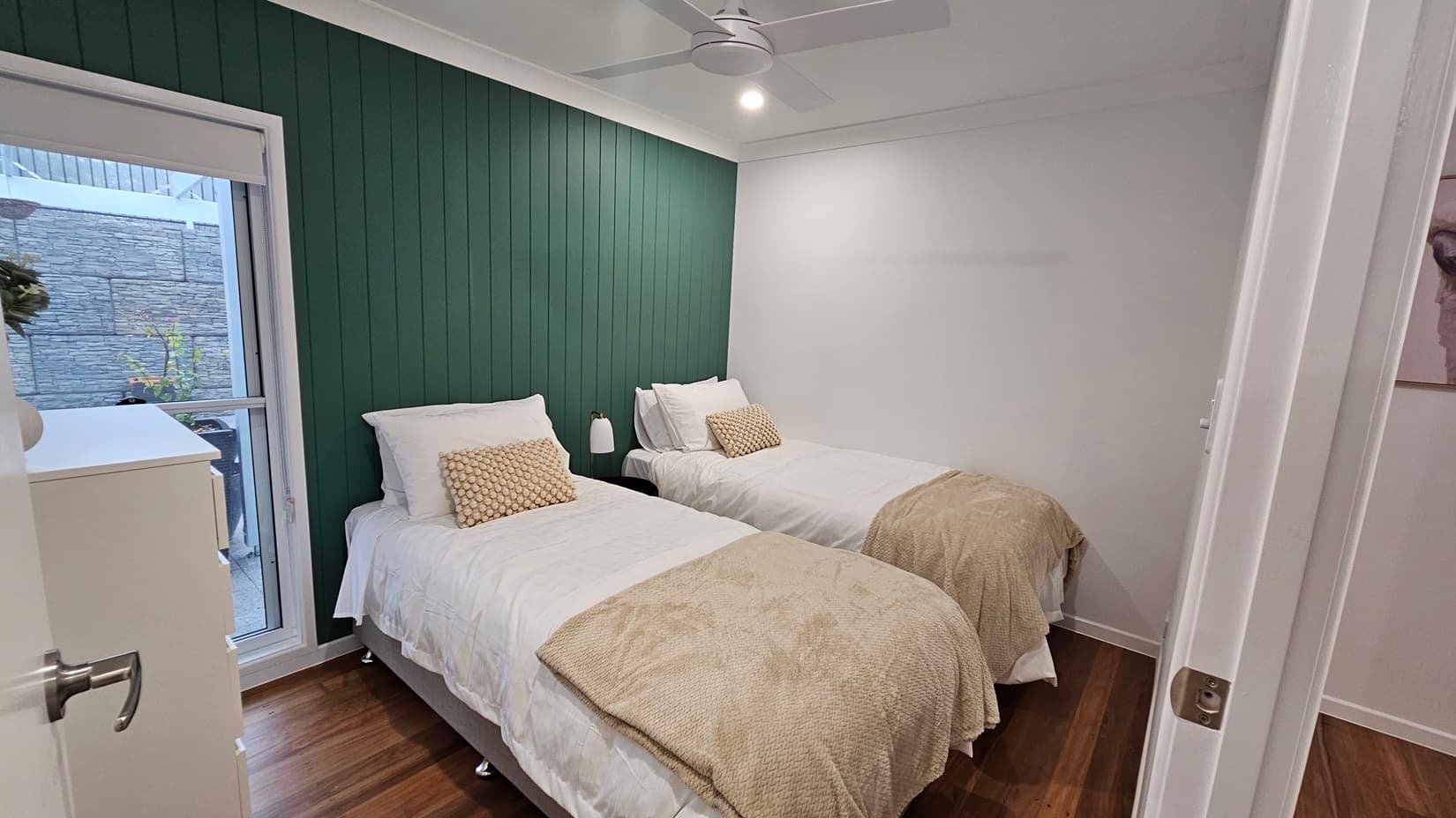 On the left, there is a green panelled wall with a window and a white chest of drawers. In the middle, two single beds with white bedding and beige throws, each with a beige cushion. On the right, a white door is slightly open, revealing part of an adjacent room. The floor is wooden, and the ceiling is white with a ceiling fan and recessed lighting.