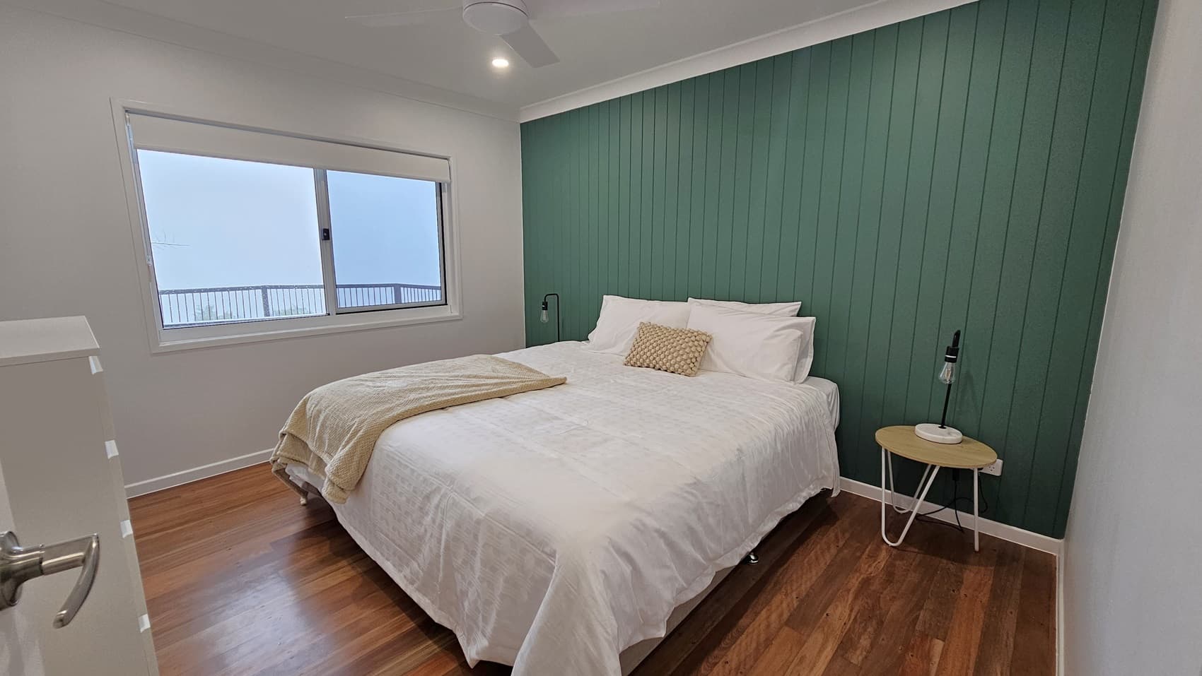 On the left, there is a white chest of drawers and a large window with a white blind. In the middle, a double bed with white bedding, a beige throw, and a beige cushion. On the right, a small round wooden side table with a black lamp sits against a green panelled wall. The floor is wooden, and the ceiling is white with a ceiling fan and recessed lighting.