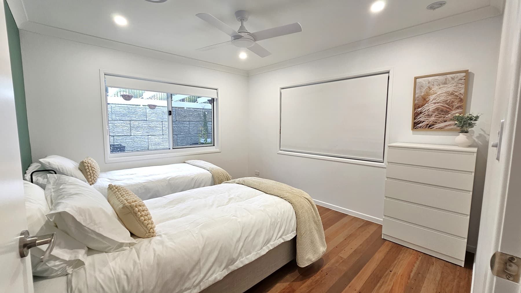 On the left, there are two single beds with white bedding, beige throws, and beige cushions, positioned under a large window with a white blind. On the right, a white chest of drawers with a framed picture above it and a small potted plant on top. The floor is wooden, and the ceiling is white with recessed lighting and a ceiling fan.