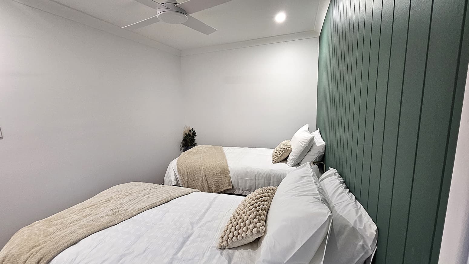 On the left, there is a plain white wall. In the middle, two single beds with white bedding, beige throws, and beige cushions are positioned head-to-head. On the right, a green panelled wall runs the length of the room. The floor is not visible, and the ceiling is white with a ceiling fan and recessed lighting.
