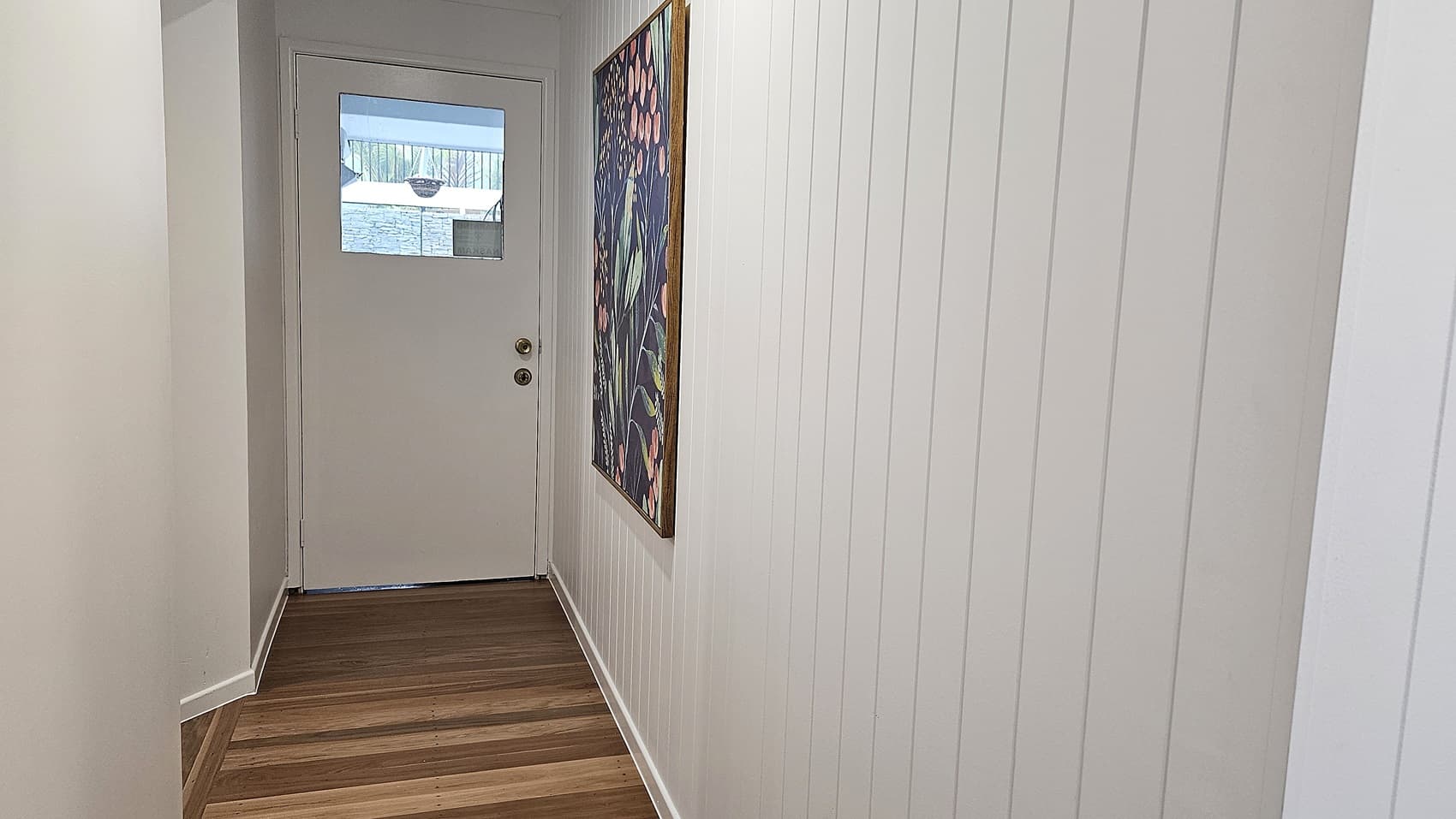 On the left, there is a plain white wall. In the middle, a white door with a small rectangular window near the top. On the right, a white panelled wall with a large colourful painting featuring abstract floral patterns. The floor is wooden with alternating light and dark planks, and the ceiling is white.